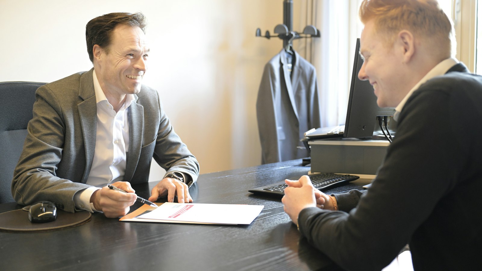 A supplier and a business customer seated across a dark office desk, smiling as they work through an unpaid invoice on a printed statement, one of them holding a pen