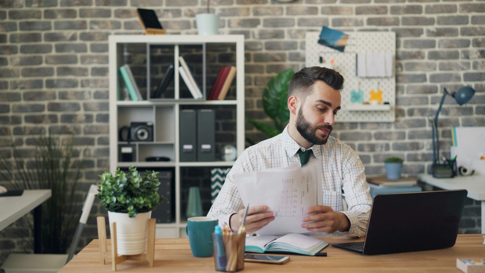 A credit manager at an office desk reading two printed invoice pages beside an open laptop while checking the paperwork on an unpaid account