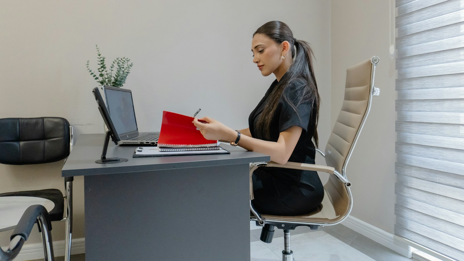 A credit manager at a grey office desk, pen in hand, checking a red account folder beside her open laptop before placing the account with a commercial collection agency